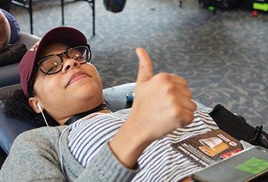 Person wearing hat, reading glasses and headphones laying on cot giving a thumbs up.