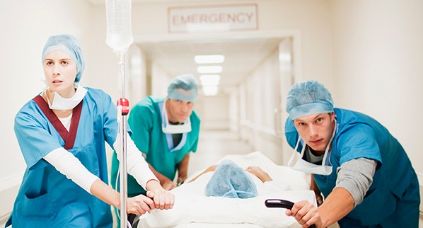 Three doctors moving patient laying in hospital bed down a hallway.
